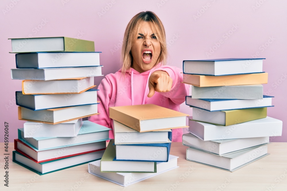 Young caucasian woman sitting on the table with books pointing displeased and frustrated to the camera, angry and furious with you Young caucasian woman sitting on the table with books pointing displeased and frustrated to the camera, angry and furious with you