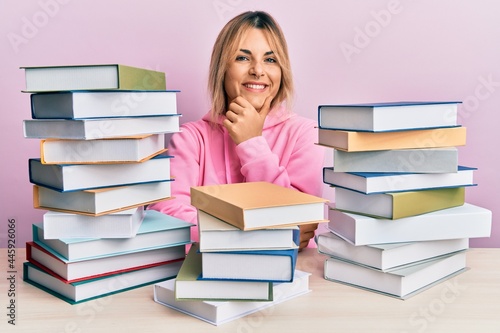 Young caucasian woman sitting on the table with books looking confident at the camera smiling with crossed arms and hand raised on chin. thinking positive.