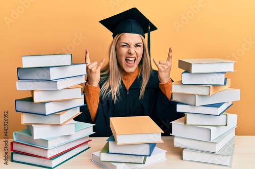 Young caucasian woman wearing graduation ceremony robe sitting on the table shouting with crazy expression doing rock symbol with hands up. music star. heavy music concept.