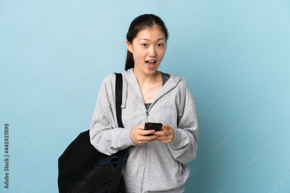 Young sport Chinese woman with sport bag over isolated blue background surprised and sending a message Young sport Chinese woman with sport bag over isolated blue background surprised and sending a message