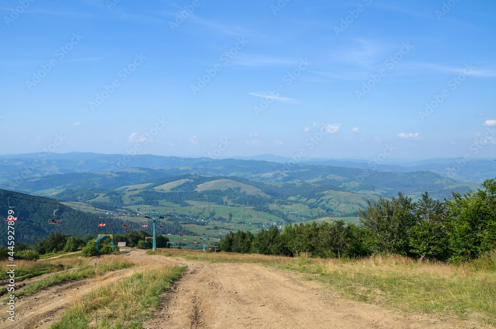 Fototapeta premium Summer mountain landscape with ski lift on the green grassy hill, and the valley with Pylypets village at sunny day. Carpathian Mountains, Ukraine
