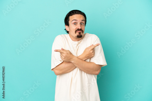 Young caucasian man isolated on blue background pointing to the laterals having doubts