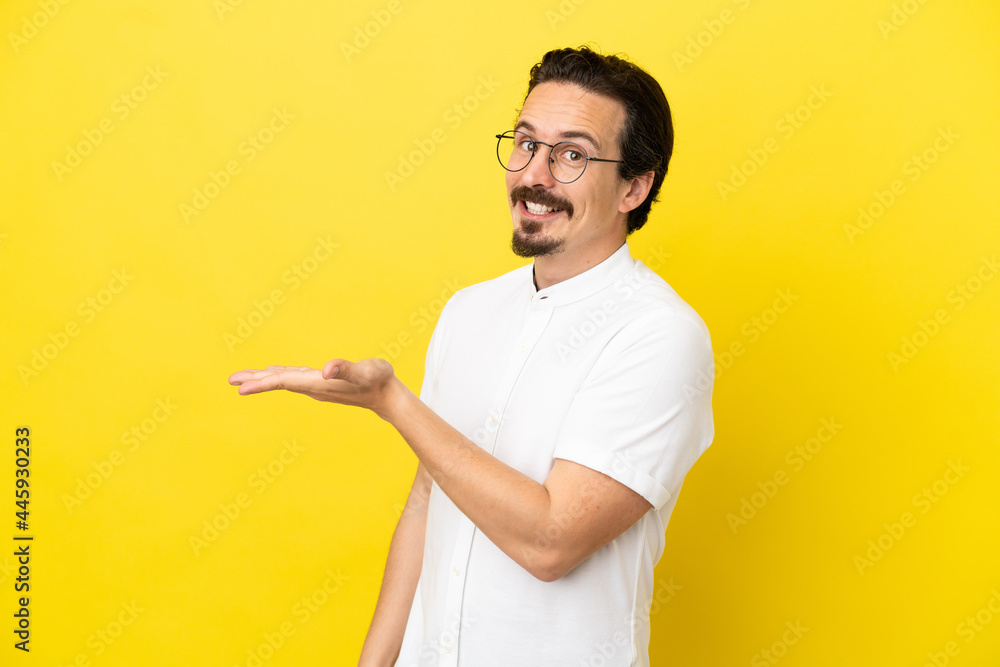 Young caucasian man isolated on yellow background presenting an idea while looking smiling towards Young caucasian man isolated on yellow background presenting an idea while looking smiling towards