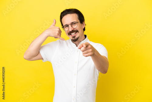Young caucasian man isolated on yellow background making phone gesture and pointing front