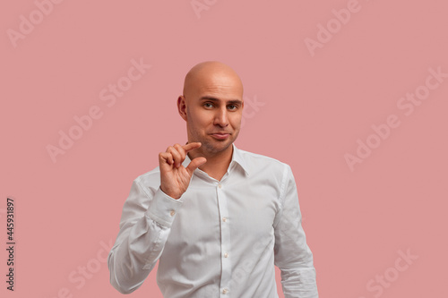 Few inches, centimeter. Funny bald man with bristle in white shirt, showing a little bit gesture, asks for something, assuring that it is just a little bit. Isolated over pink background.