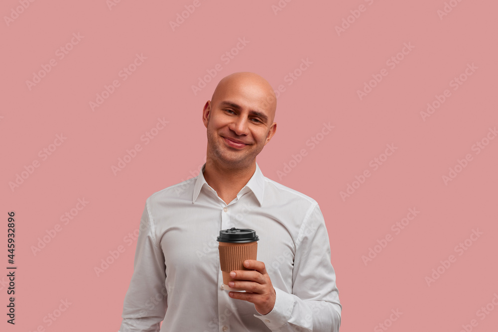 Joyful smiling bald man with bristle holds paper takeaway cup, enjoys aromatic beverage at work with colleague. Dressed in white shirt. Isolated over pink background.