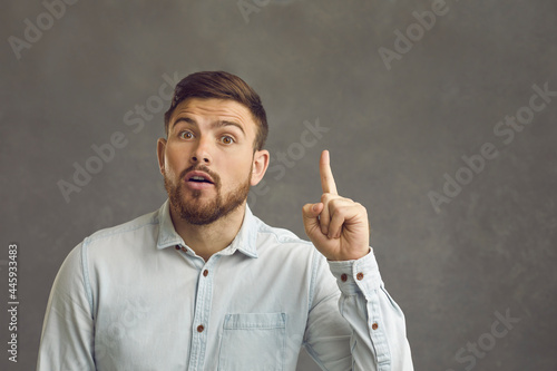 I have an idea. Positive young man raises his index finger up as a sign that some bright idea or thought has come to his mind. Handsome bearded millennial man posing in studio on gray wall background.