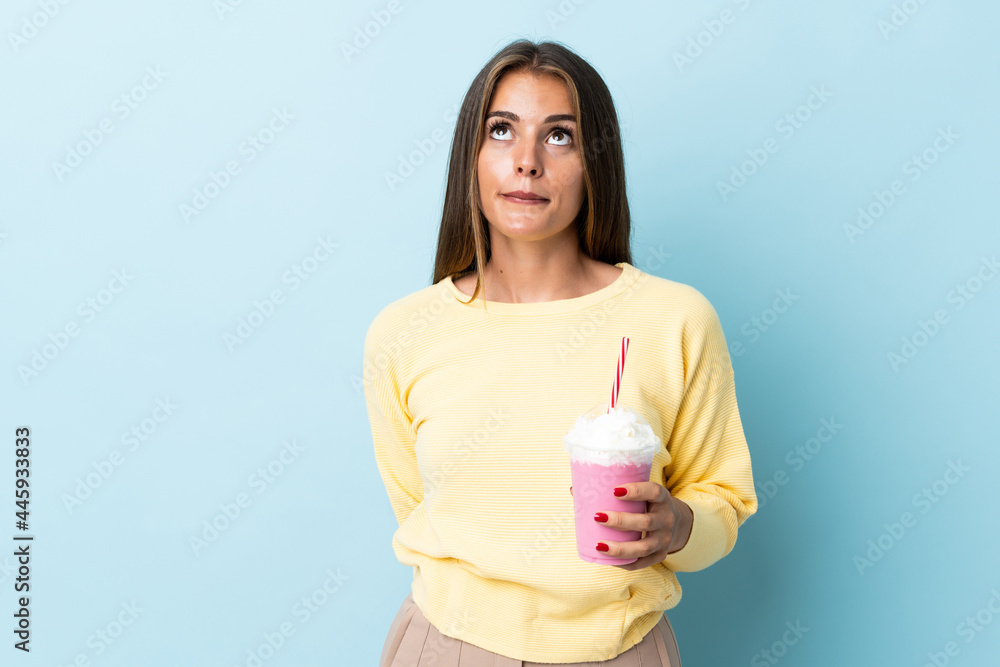 Young Uruguayan woman with strawberry milkshake isolated on blue background and looking up Young Uruguayan woman with strawberry milkshake isolated on blue background and looking up