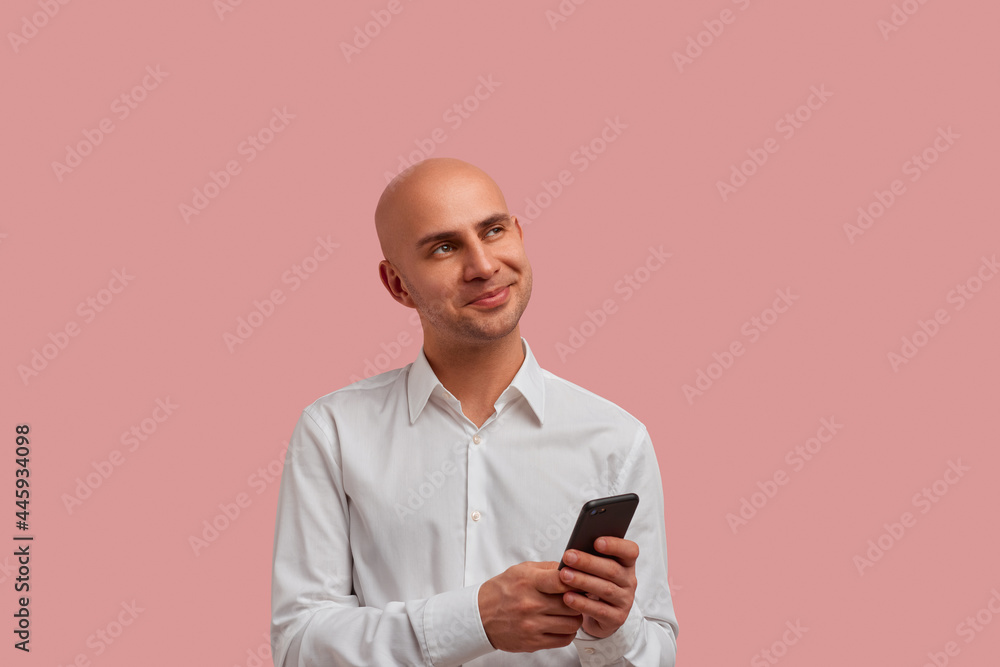 Horizontal photo shoot of thoughtful bald man holds smartphone, looks pensive. Dreaming about how he meet his pen pal from other country. Has bristle. Dressed in white shirt. Horizontal photo shoot of thoughtful bald man holds smartphone, looks pensive. Dreaming about how he meet his pen pal from other country. Has bristle. Dressed in white shirt.