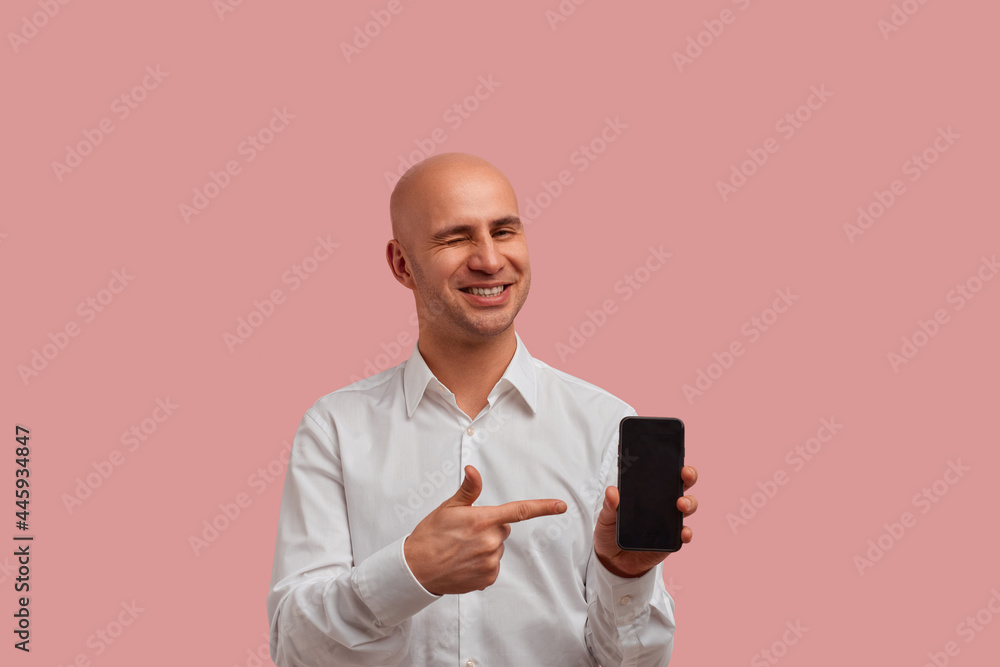 Horizontal photoshot of funny bald man with bristle in white shirt, winks and points to blank of smartphone display, recommend product. looks happy, glad to help you. Isolated on pink background. Horizontal photoshot of funny bald man with bristle in white shirt, winks and points to blank of smartphone display, recommend product. looks happy, glad to help you. Isolated on pink background.