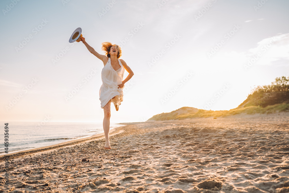 Happy young woman wearing white long dress and hat dance and jump on beach during dawn. Happiness, freedom