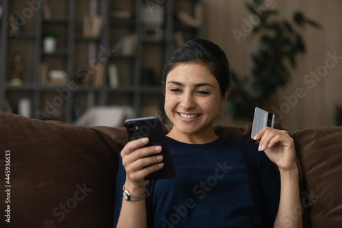 Smiling young Indian female buyer client shopping online on smartphone with credit debit card. Happy mixed race woman use cellphone make payment buy on internet on gadget. Consumerism concept.