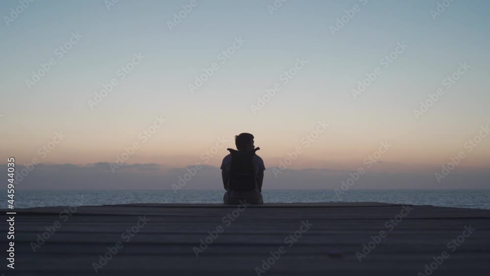 Theme of loneliness, depression, search for answers to questions while traveling. Man traveler with backpack looks at sunset of Mediterranean sea from wooden pier on island of Cyprus, city of Paphos. Theme of loneliness, depression, search for answers to questions while traveling. Man traveler with backpack looks at sunset of Mediterranean sea from wooden pier on island of Cyprus, city of Paphos.