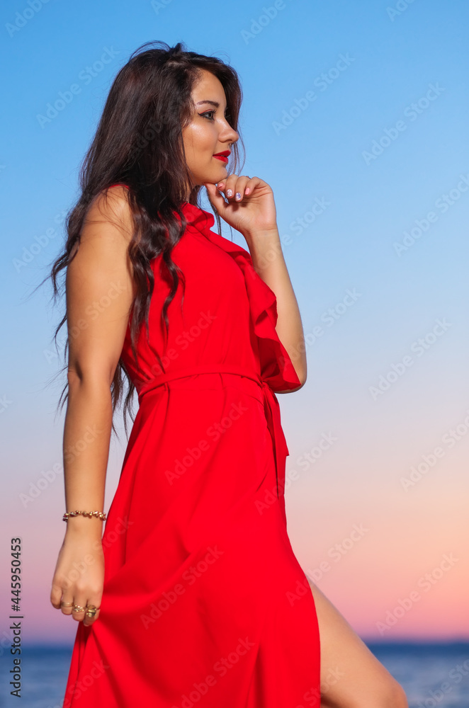 Beautiful brunette girl in a red dress posing on the seashore during sunrise or sunset