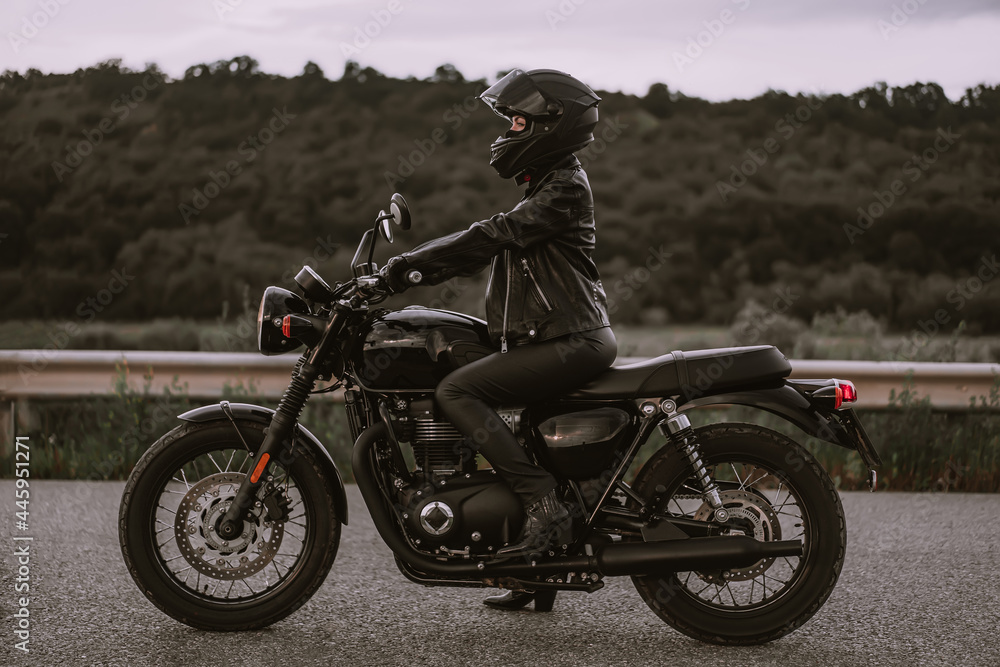 Portrait of confident motorcyclist woman in motorcycle helmet. Young driver biker looking away outdoors alone on highway. Ready for trip. Cafe racers, motorbike aesthetics and vintage design concept.