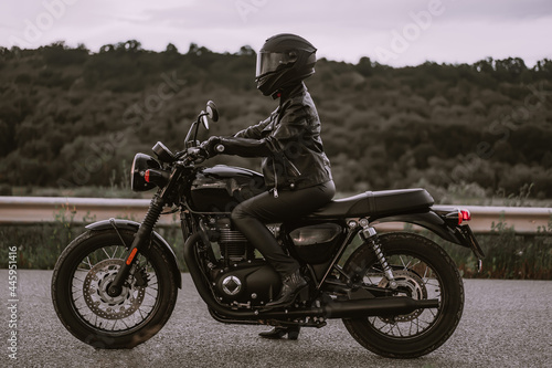 Portrait of confident motorcyclist woman in motorcycle helmet. Young driver biker looking away outdoors alone on highway. Ready for trip. Cafe racers, motorbike aesthetics and vintage design concept.