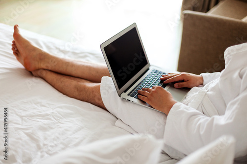 a man in a robe lies on a bed with a computer on his feet. Freelance, work from home. No face. A man in a hotel, working in a bedroom at a computer.