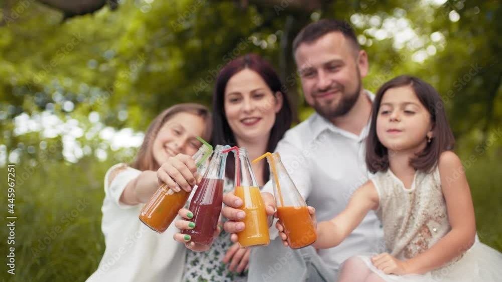 Happy young family with two children drinking fresh juice while sitting on blanket at green garden. Caucasian parents with two cute daughters drinking juice in glass bottle from colorful straws Happy young family with two children drinking fresh juice while sitting on blanket at green garden. Caucasian parents with two cute daughters drinking juice in glass bottle from colorful straws