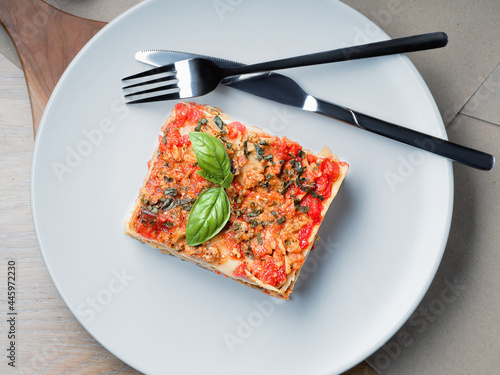 Piece of lasagna. White plate with fork and knife. Basil leafs, tomatoes and zucchini on table. Menu of Italian cuisine. Top down view