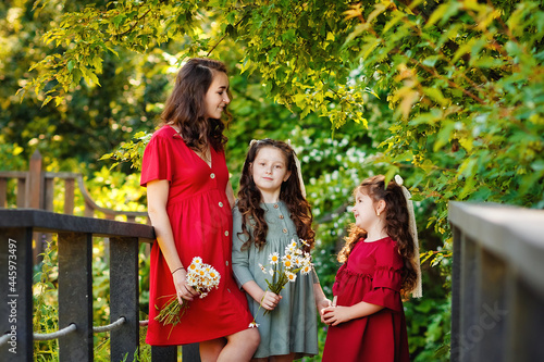 A pregnant mother in a red dress and her two daughters in nature. The family is standing on the bridge holding a bouquet of daisies in their hands.