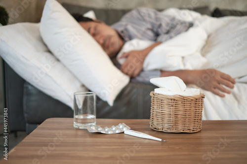 Cuadro en lienzo Asian man is absent from work and sick leave because of illness, takes temperature by thermometer, takes medicine, and sleeps in blankets on sofa at home, tissues, and capsule pills on the table