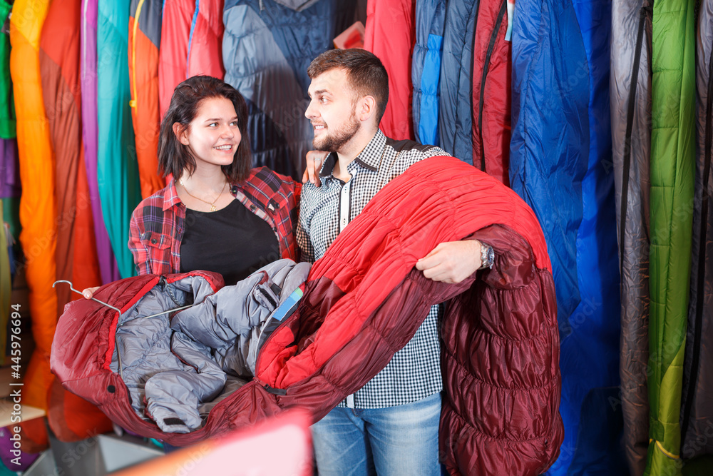 Cheerful couple examining various sleeping bags in sports equipment store
