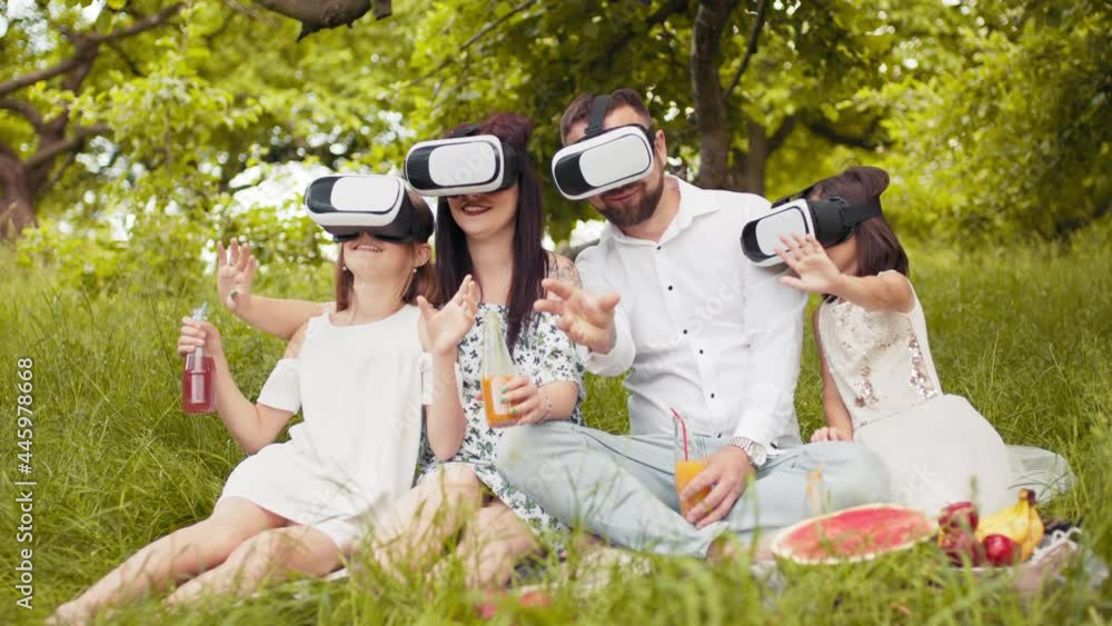 Young caucasian family having fun in virtual glasses on fresh air. Happy parents with two daughters sitting on soft blanket and playing games. Leisure time and entertainment concept. Happy family Young caucasian family having fun in virtual glasses on fresh air. Happy parents with two daughters sitting on soft blanket and playing games. Leisure time and entertainment concept. Happy family