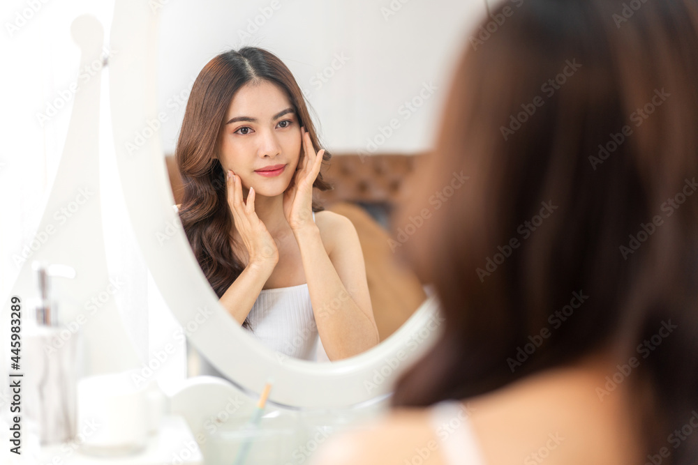 Smiling of young beautiful pretty asian woman clean fresh healthy white skin looking at mirror.asian girl touching on her face with hand and applying cream at home.spa and beauty concept Smiling of young beautiful pretty asian woman clean fresh healthy white skin looking at mirror.asian girl touching on her face with hand and applying cream at home.spa and beauty concept