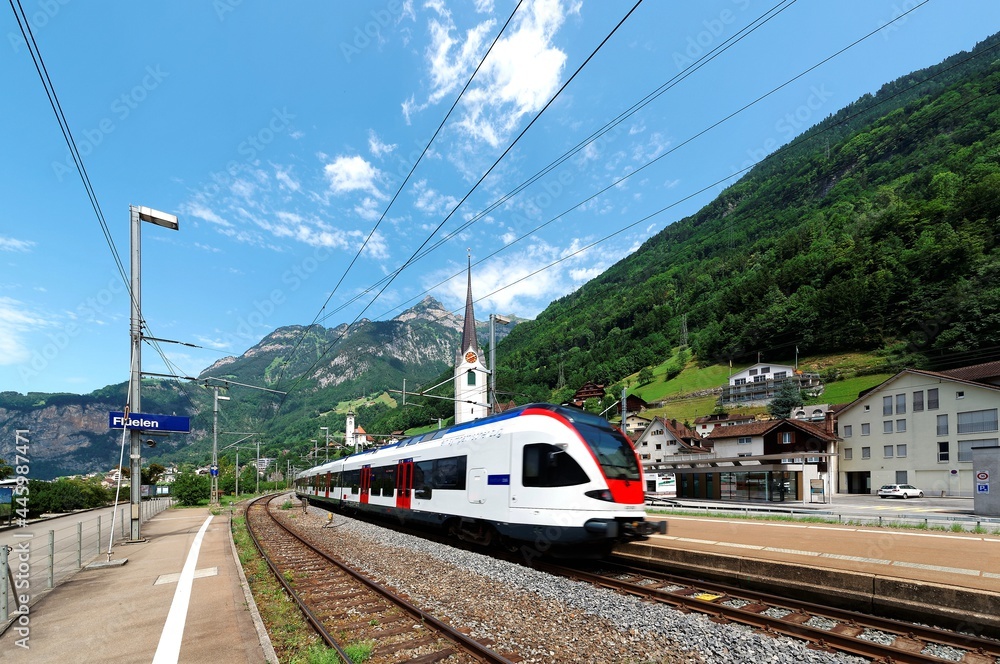 A train travels through a railway curve in beautiful Fluelen Village on a sunny summer day, with churches on the hillside under majestic Swiss Alpine mountains in the background, Switzerland, Europe A train travels through a railway curve in beautiful Fluelen Village on a sunny summer day, with churches on the hillside under majestic Swiss Alpine mountains in the background, Switzerland, Europe