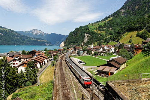 Scenery of beautiful Lake Lucerne on a sunny summer day, with a train traveling on the railway thru Sisikon Village by Lake Uri & majestic mountains under blue clear sky in background, in Switzerland photo