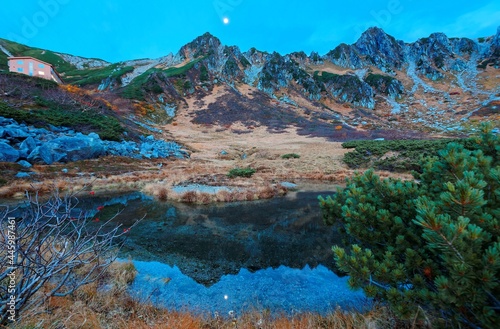 Fall scenery of Senjojiki Cirque in morning twilight with rugged Kiso mountain peaks in background reflected in a pond & the moon rising in the sky in Japanese Central Alps National Park, Nagano Japan