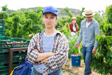 Portrait of confident woman farmer posing in fruit garden while team of workers picking sweet cherry