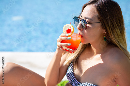 young woman sipping a refreshing drink by the side of the pool wearing sunglasses on summertime