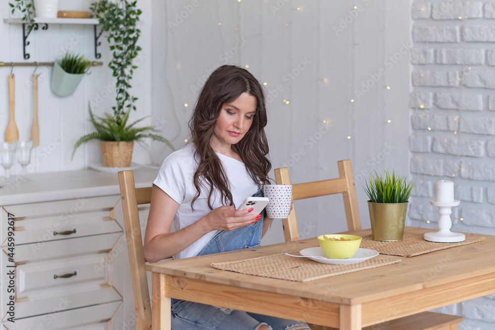 Young beautiful woman using cell phone and having a coffee in the kitchen. Young beautiful woman using cell phone and having a coffee in the kitchen.