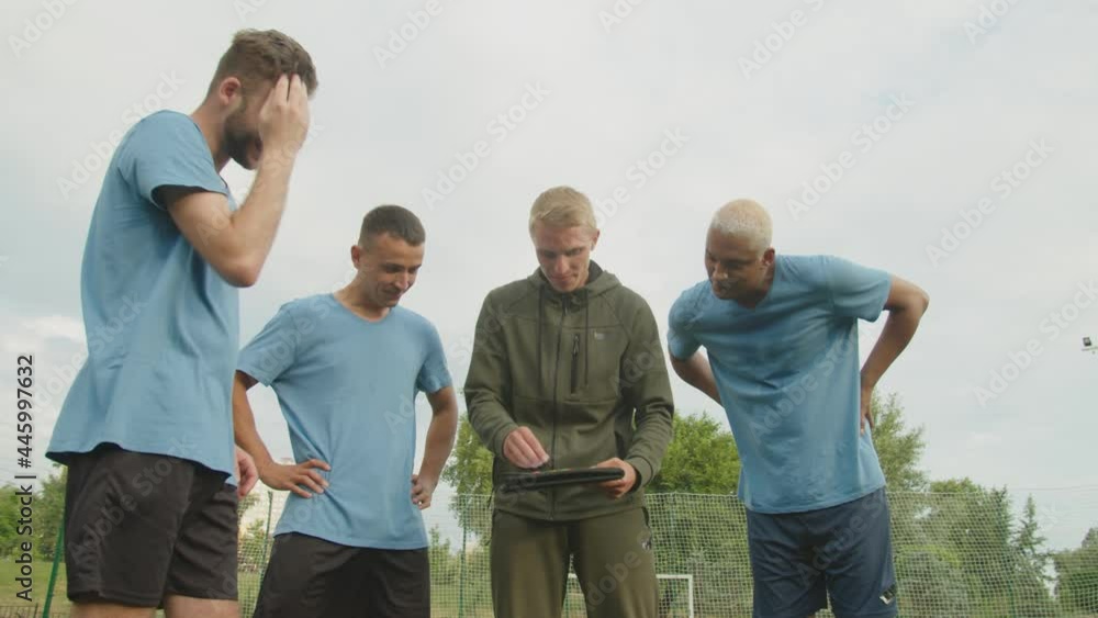 Low section of handsome football coach using tactic board, explaining scheme of game match to group of diverse multiethnic cheerful sportsmen in football sportswear outside at daybreak Low section of handsome football coach using tactic board, explaining scheme of game match to group of diverse multiethnic cheerful sportsmen in football sportswear outside at daybreak