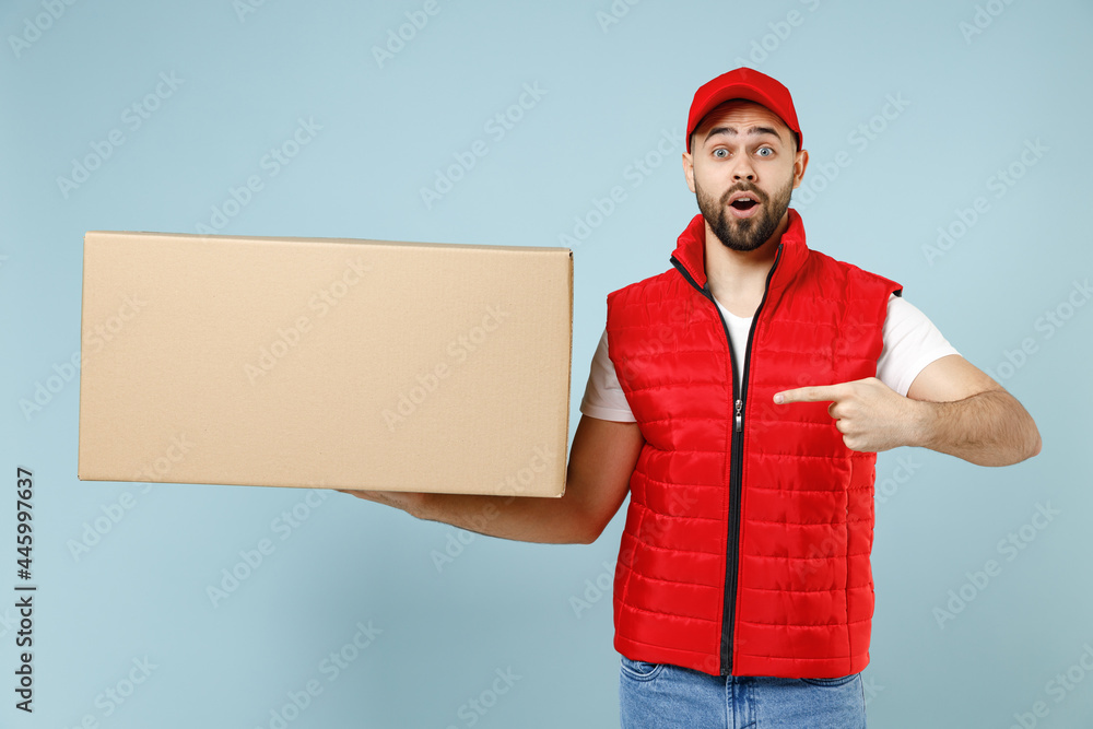 Professional delivery guy employee man in red cap white T-shirt vest uniform workwear work as dealer courier hold blank cardboard box isolated on pastel blue color background studio. Service concept. Professional delivery guy employee man in red cap white T-shirt vest uniform workwear work as dealer courier hold blank cardboard box isolated on pastel blue color background studio. Service concept.