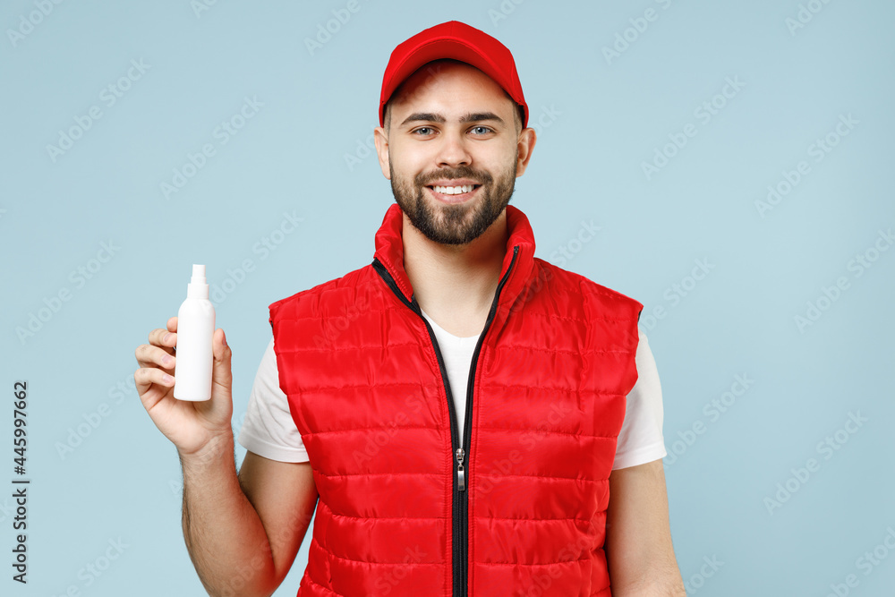 Delivery guy employee man in red cap white T-shirt vest uniform workwear work dealer courier hold bottle spray sanitizer soap isolated on pastel blue color background studio portrait. Service concept. Delivery guy employee man in red cap white T-shirt vest uniform workwear work dealer courier hold bottle spray sanitizer soap isolated on pastel blue color background studio portrait. Service concept.