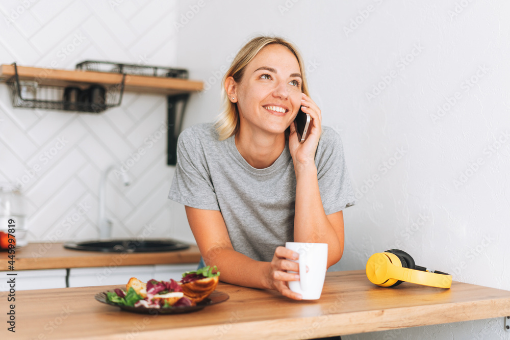 Young smiling blonde woman in yellow t-shirt with cup of tea talking on mobile phone in hands in the kitchen at home