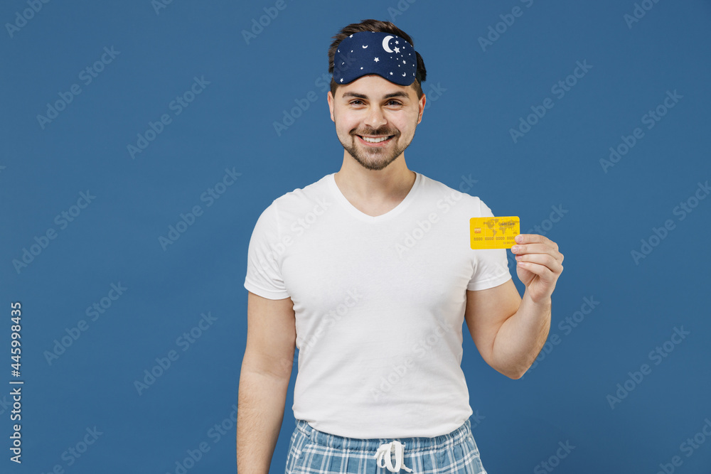 Young smiling rich fun calm caucasian man 20s wearing pajama jam sleep mask rest relax at home holding in hand credit bank card isolated on dark blue background studio Good mood night bedtime concept Young smiling rich fun calm caucasian man 20s wearing pajama jam sleep mask rest relax at home holding in hand credit bank card isolated on dark blue background studio Good mood night bedtime concept