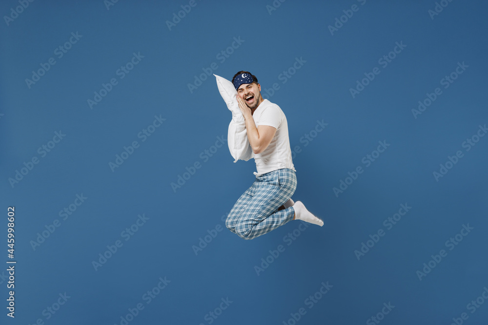 Full length side view fun young caucasian man 20s wearing pajamas jam sleep mask resting relax at home jump high lie on pillow scream isolated on dark blue background. Good mood night bedtime concept Full length side view fun young caucasian man 20s wearing pajamas jam sleep mask resting relax at home jump high lie on pillow scream isolated on dark blue background. Good mood night bedtime concept