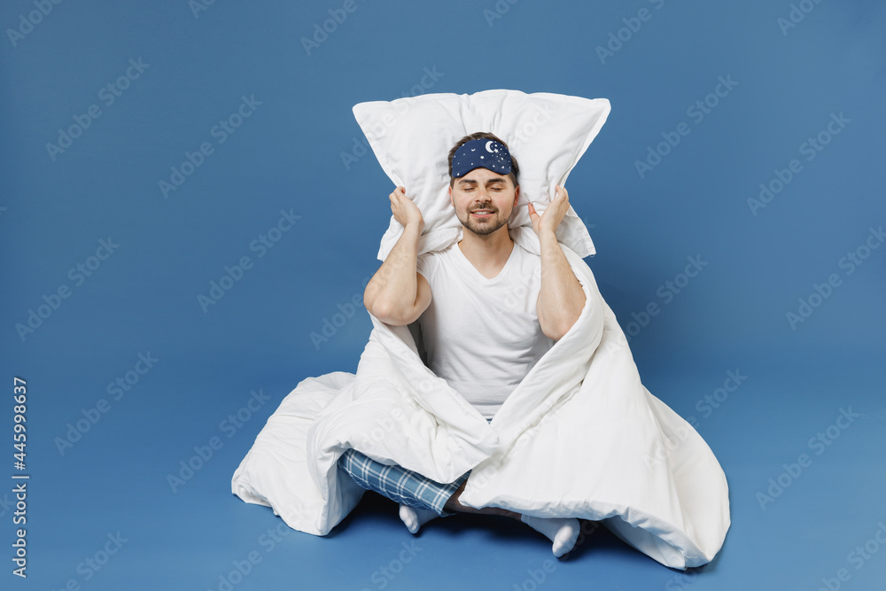 Full size body length calm young man in pajamas jam sleep mask rest relax at home cover ears with pillow sit wrap cover blanket duvet isolated on dark blue background Bad mood night bedtime concept. Full size body length calm young man in pajamas jam sleep mask rest relax at home cover ears with pillow sit wrap cover blanket duvet isolated on dark blue background Bad mood night bedtime concept.