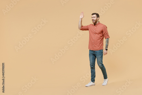 Full size body length happy tatooed young brunet man 20s short haircut wears apricot shirt stride meet greet wave hand as notices someone strength isolated on pastel orange background studio portrait.