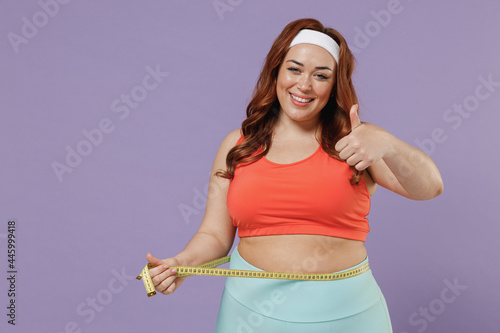 Young satisfied happy chubby overweight plus size big fat fit woman wear red top hold measuring tape on waist show thumb up warm up train isolated on purple background gym home. Workout sport concept. photo