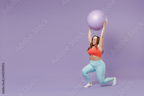 Full length side view young chubby overweight plus size big fat fit woman wearing red top warm up training squats with fit ball raised up hands isolated on purple background gym Workout sport concept © ViDi Studio