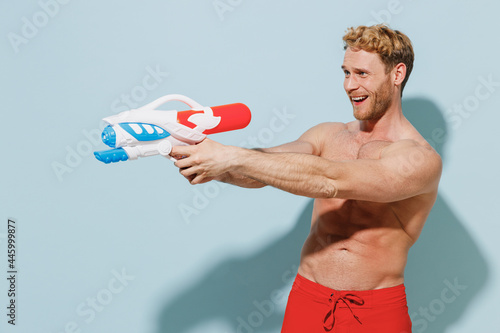 Side view handsome young fun sexy man in red shorts swimsuit relax near hotel pool hold in hand play shoot from water gun isolated on pastel blue background. Summer vacation sea rest sun tan concept