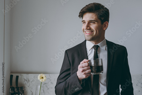 Young smiling minded successful employee business man corporate lawyer 20s wearing formal black suit shirt tie drink coffee breakfast look aside in light kitchen Achievement career lifestyle concept.