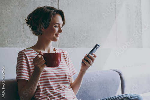 Young caucasian smiling happy woman 20s in casual clothes sitting on grey sofa using mobile cell phone hold red cup drink coffee look aside rest indoors flat at home People lifestyle leisure concept