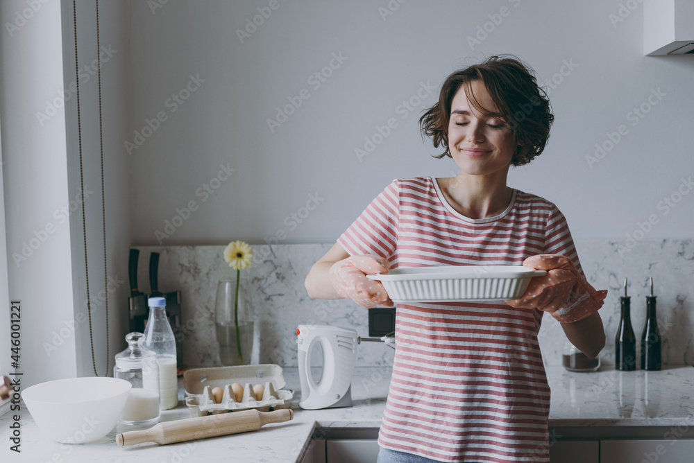 Young housewife woman 20s in casual clothes striped tshirt holding hot casserole in baking dish sniff delicious scent cooking food in light kitchen at home alone Healthy diet bakery lifestyle concept Young housewife woman 20s in casual clothes striped tshirt holding hot casserole in baking dish sniff delicious scent cooking food in light kitchen at home alone Healthy diet bakery lifestyle concept