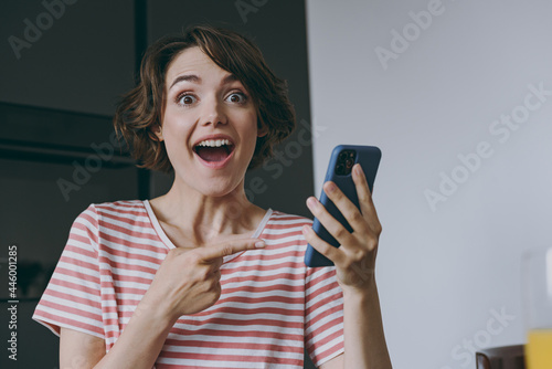 Young surprised housewife woman in casual clothes striped t-shirt point index finger on mobile cell phone chatting online browsing internet in light kitchen at home alone People lifestyle concept. photo