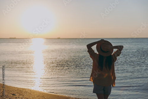 Silhouette back rear view young stylish woman 20s wearing straw hat shirt summer casual clothes standing resting outdoors at sunrise sun dawn over sea beach. People vacation lifestyle journey concept.